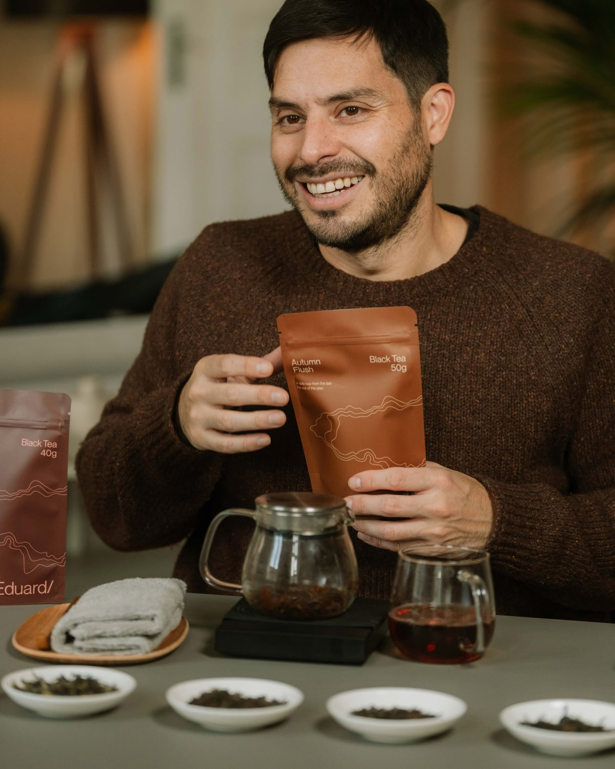 Eduardo smiling at a tasting table with a pouch, a glass teapot and tea leaves